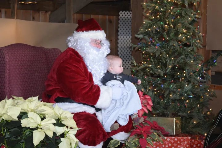 Santa holds a baby on his lap next to a decorated Christmas tree and poinsettias.