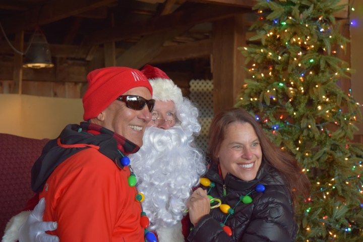 Two people smiling with Santa Claus, standing near a decorated Christmas tree.