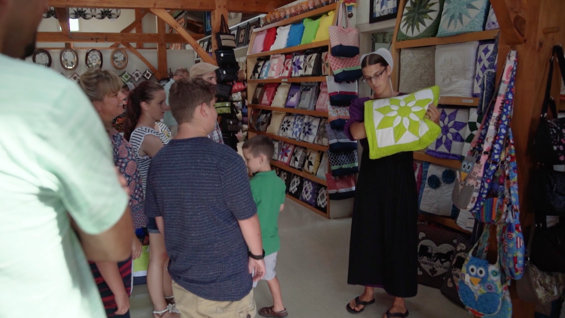 Woman shows green quilt to group in a shop with quilts and bags on shelves.