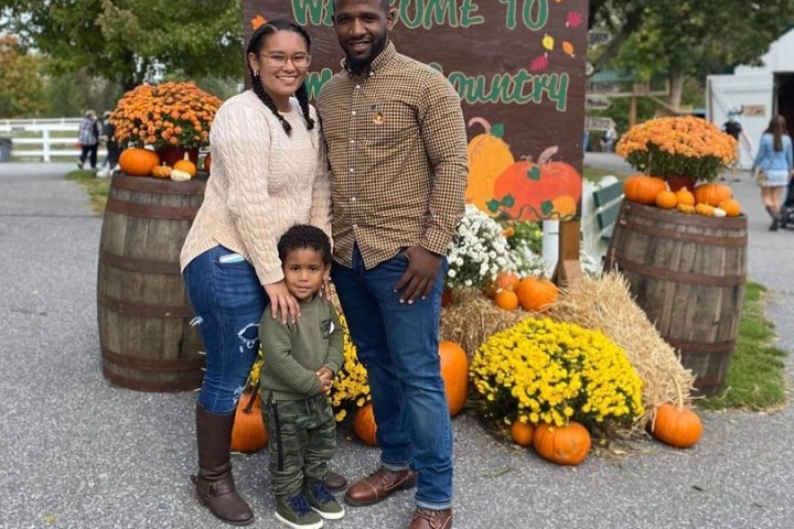 Family of three posing in front of fall-themed decorations with pumpkins and flowers.