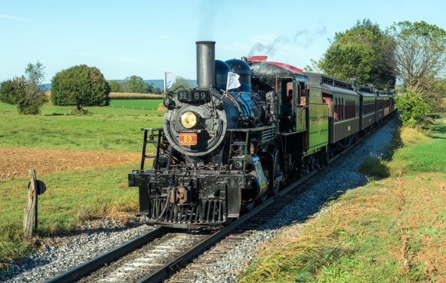 Steam train traveling on a rural track with green fields on either side.